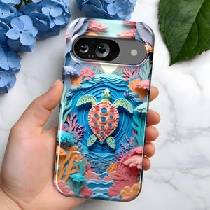 May include: A phone case featuring a 3D-style design of a sea turtle swimming in a coral reef. The case has a clear border and a colorful design with shades of blue, pink, and orange. The phone case is held in a hand.