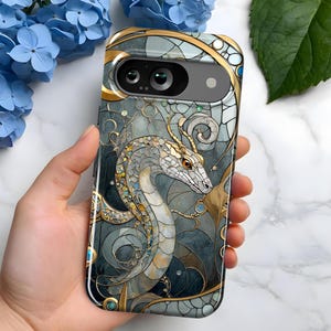May include: A phone case featuring a stained-glass style dragon design. The case has a gold and teal color scheme with a detailed illustration of a dragon. The dragon is white and gold, with a long, serpentine body. The case is held in a hand.