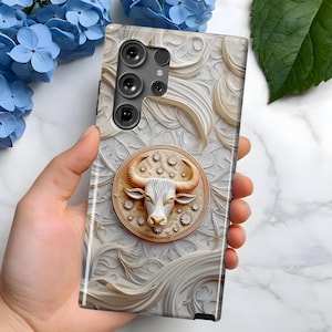 May include: A white phone case with a gold and white design featuring a bull's head in a circle. The phone case has a camera lens cutout and a white background.