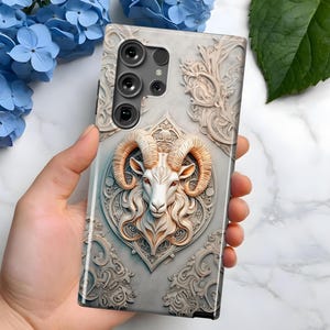 May include: A white phone case featuring an intricate design of a ram's head within a decorative frame. The case has a camera lens cutout with three lenses.