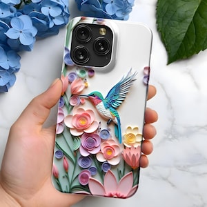 May include: A white phone case featuring a colorful hummingbird and floral design. The case has a 3D effect with pink, purple, and yellow flowers. The hummingbird has blue, green, and red feathers. The case is held in a hand.