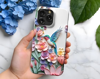 May include: A white phone case featuring a colorful hummingbird and floral design. The case has a 3D effect with pink, purple, and yellow flowers. The hummingbird has blue, green, and red feathers. The case is held in a hand.