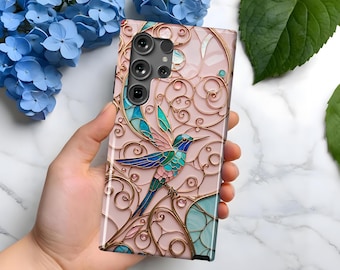 May include: A pink phone case with a gold and blue hummingbird design. The hummingbird is surrounded by intricate gold swirls and the phone case has a camera cutout.