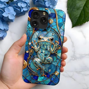 Puede incluir: Una funda de teléfono azul y dorada con un diseño de mosaico que presenta una rana. La funda tiene un recorte para las lentes de la cámara del teléfono.
