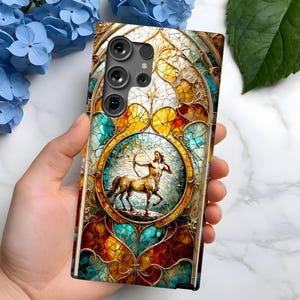 May include: A phone case with a stained glass design featuring a mythical creature with a bow and arrow. The case has a gold frame and a camera lens cutout.