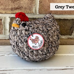 Puede incluir: Un pollo de crochet de tweed gris con un sombrero rojo y una etiqueta que dice "Merry Chickmas".