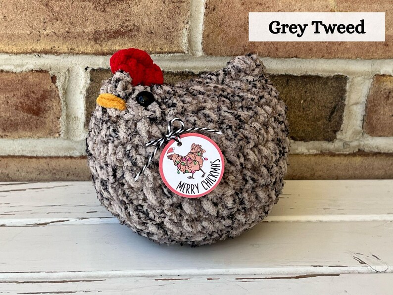 Puede incluir: Un pollo de crochet de tweed gris con un sombrero rojo y una etiqueta que dice "Merry Chickmas".