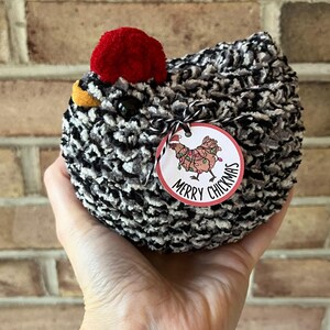 Puede incluir: Un juguete de peluche de pollo negro y blanco con un pomp&oacute;n rojo en la cabeza. El juguete tiene una etiqueta de regalo adjunta que dice "Merry Chickmas" con un pollo de dibujos animados que lleva luces de Navidad.