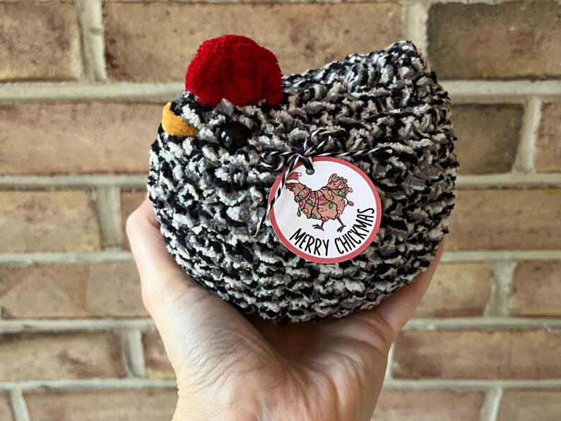 Puede incluir: Un juguete de peluche de pollo negro y blanco con un pomp&oacute;n rojo en la cabeza. El juguete tiene una etiqueta de regalo adjunta que dice "Merry Chickmas" con un pollo de dibujos animados que lleva luces de Navidad.