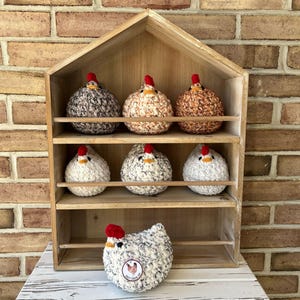 Puede incluir: Una estanter&iacute;a de madera con siete pollos de crochet en varios colores y patrones. Los pollos est&aacute;n dispuestos en tres estantes, con un pollo sentado en el estante inferior frente a la unidad.