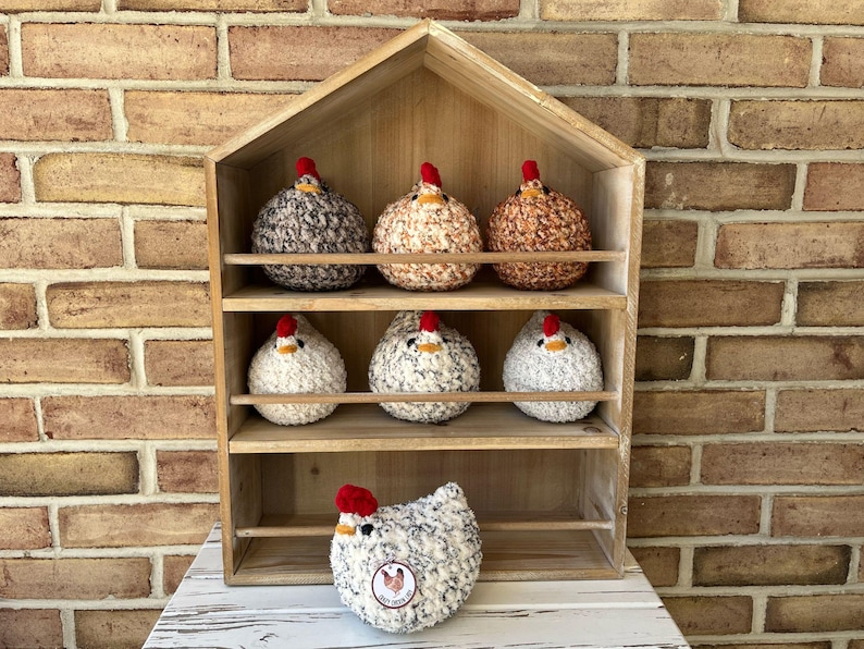 Puede incluir: Una estanter&iacute;a de madera con siete pollos de crochet en varios colores y patrones. Los pollos est&aacute;n dispuestos en tres estantes, con un pollo sentado en el estante inferior frente a la unidad.