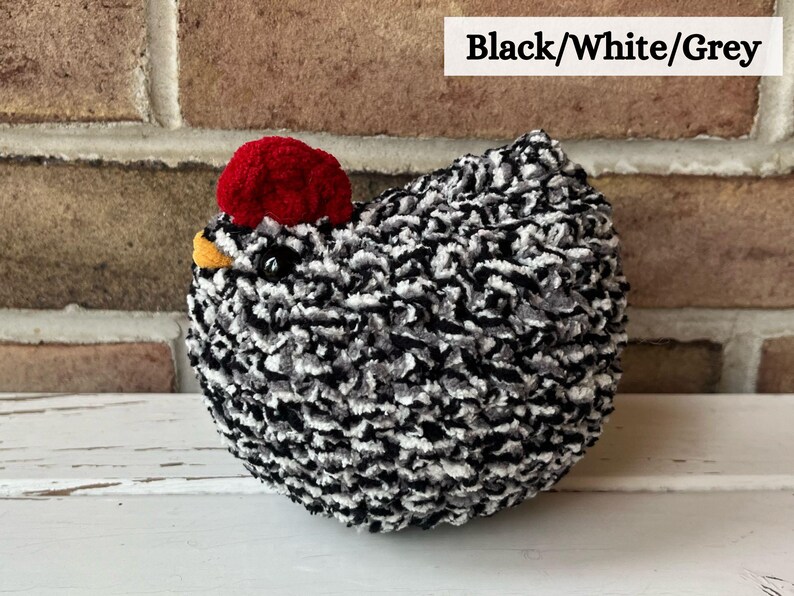 Puede incluir: Juguete de pollo de peluche negro, blanco y gris con una cresta roja. El pollo est&aacute; sentado sobre una superficie blanca.
