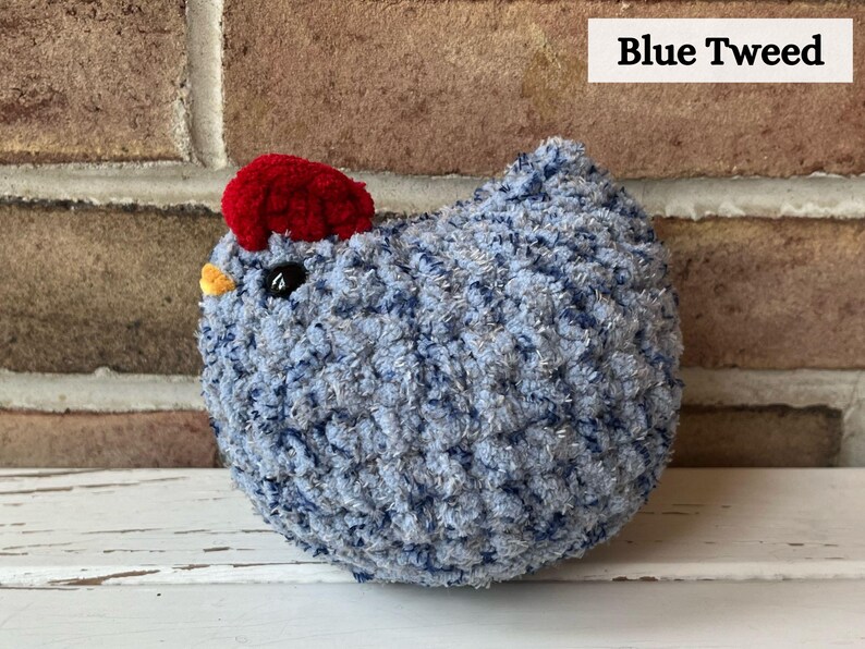 Puede incluir: Un pollo de crochet azul y plateado con una cresta de fieltro rojo. El pollo est&aacute; sentado sobre una superficie blanca. Blue Tweed.