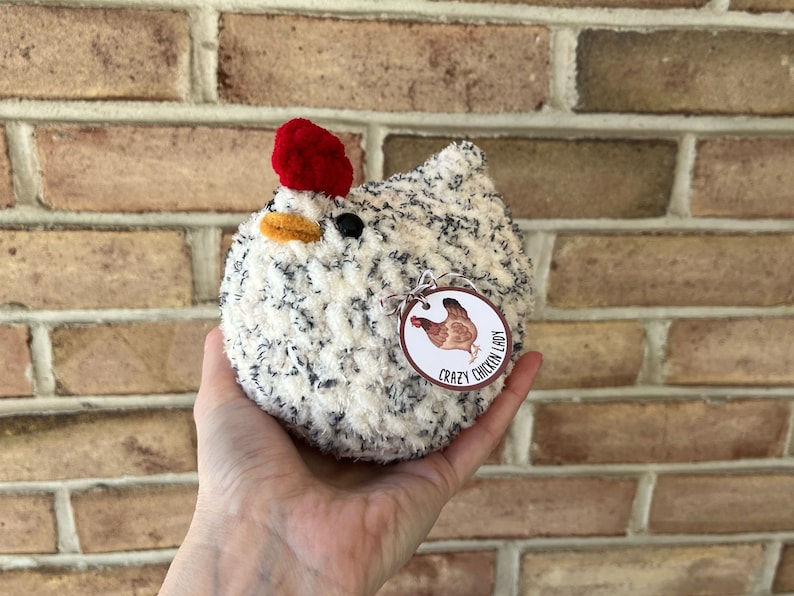 Puede incluir: Un juguete de pollo de crochet blanco y negro con una cresta roja y una etiqueta que dice "Crazy Chicken Lady".