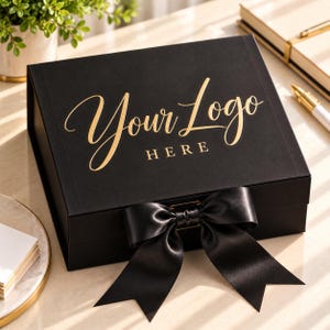 May include: A rectangular black gift box featuring the gold foil text Your Logo HERE on the lid. The box is secured with a wide black satin ribbon tied into a bow at the front center, creating a sophisticated presentation for various items.
