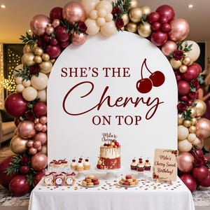 May include: A white arched backdrop featuring the text "SHE'S THE Cherry ON TOP" with a cherry graphic, surrounded by a balloon garland in burgundy, rose gold, and cream. A table holds a cake, cupcakes, macarons, and a sign reading "Mila's Cherry Sweet Birthday!"