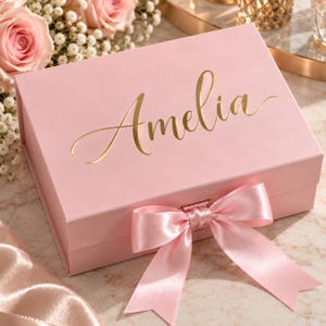 May include: A rectangular light pink gift box featuring the name Amelia written in elegant gold cursive script on the lid. The box is secured with a matching light pink satin ribbon tied into a bow at the front center, creating a clean look.