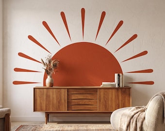 May include: A large, semi-circular terracotta-colored sun wall decal featuring radiating rays. The graphic is applied to a plain off-white wall, positioned directly above a wooden sideboard that holds a small vase with dried botanicals and books.