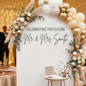 Può includere: Scena di festa di fidanzamento con un arco bianco decorato con palloncini, fiori ed erba della pampa. L'arco presenta il testo "Celebrating the Future Mr. & Mrs. Smith". Un tavolino espone un libro degli ospiti e un cartello.