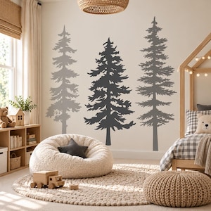 Può includere: Tre grandi adesivi murali in vinile grigio scuro che raffigurano pini sempreverdi sono applicati su una parete chiara. Gli alberi variano in altezza e densità dei rami, creando una silhouette boschiva nella stanza.