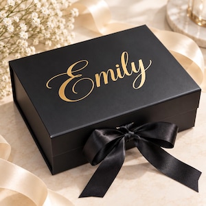 May include: A rectangular black gift box featuring the name Emily written in gold cursive script on the lid. The box is secured with a black satin ribbon tied into a bow at the front, providing a contrast against the matte black surface.
