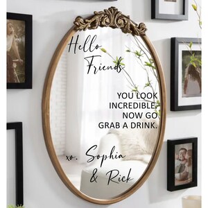May include: A gold-framed oval mirror with a handwritten message that reads "Hello Friends You Look Incredible Now Go Grab A Drink. xo. Sophia & Rick".