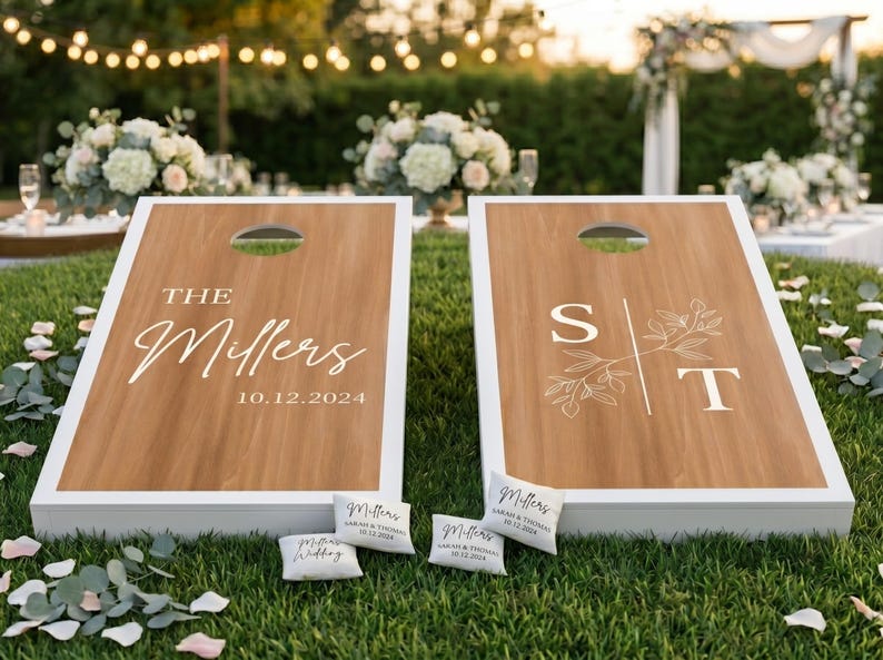 K&ouml;nnte beinhalten: Zwei personalisierte Cornhole-Boards mit wei&szlig;em Rand und Holzmaserung. Ein Board zeigt "The Millers" und das Datum "10.12.2024". Das andere Board hat die Initialen "S" und "T" mit floralen Akzenten. Kleine personalisierte Kissen liegen herum.