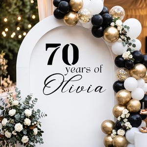 May include: A white arched backdrop featuring the black text 70 years of Olivia. The arch is decorated with a balloon garland in black, white, and metallic gold, accented with small white flowers and green eucalyptus leaves.