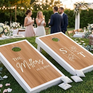 K&ouml;nnte beinhalten: Zwei individuelle Cornhole-Boards mit wei&szlig;en Rahmen und Holzoberfl&auml;chen. Ein Board zeigt "THE Millers" und das Datum "10.12.2024". Das andere Board zeigt die Initialen "S T" mit floralen Akzenten. Kleine wei&szlig;e Beanbags liegen daneben.