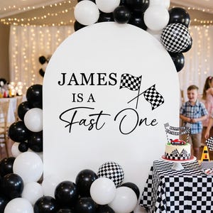 May include: A white arched backdrop featuring the black text "JAMES IS A Fast One" with two checkered racing flags. The display is decorated with a cluster of black, white, and checkered balloons, and a birthday cake with a racing theme.