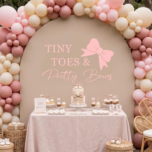 May include: A beige arched backdrop featuring the text "TINY TOES & Pretty Bows" and a pink bow graphic, surrounded by a balloon arch in shades of pink, cream, and mauve. A table with a cake, cupcakes, and treats sits in front of the backdrop.