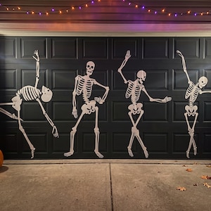 May include: Four white skeleton decals in various dancing poses are applied to a dark gray garage door. Each skeleton features detailed rib cages, skulls, and limbs arranged in dynamic positions. Two carved jack-o'-lanterns sit on the ground.