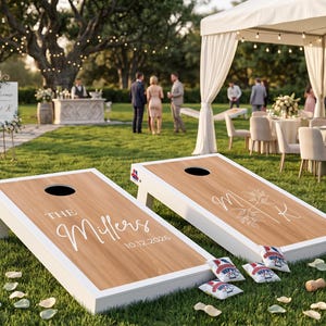 K&ouml;nnte beinhalten: Zwei individuelle Cornhole-Boards mit wei&szlig;en Rahmen und Holzmaserung stehen auf einer Rasenfl&auml;che. Auf einem Board steht "The Millers" und das Datum "10.12.2026". Das andere Board hat die Initialen "M & K". Mehrere Beanbags liegen in der N&auml;he.