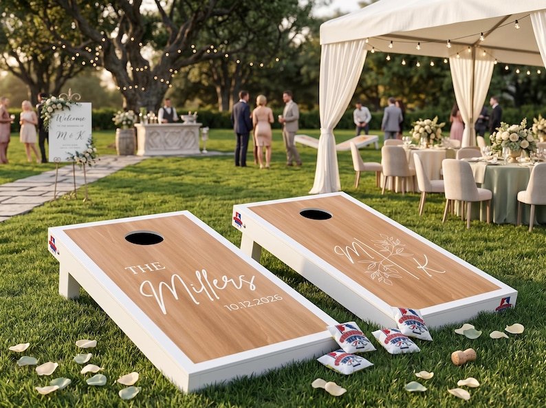 K&ouml;nnte beinhalten: Zwei individuelle Cornhole-Boards mit wei&szlig;en Rahmen und Holzmaserung stehen auf einer Rasenfl&auml;che. Auf einem Board steht "The Millers" und das Datum "10.12.2026". Das andere Board hat die Initialen "M & K". Mehrere Beanbags liegen in der N&auml;he.