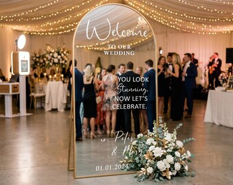 Könnte beinhalten: Ein großer, bogenförmiger goldgerahmter Spiegel mit der Aufschrift "Welcome to our Wedding" und anderem Text. Der Spiegel spiegelt eine Hochzeitsfeier mit Gästen und Blumenschmuck wider. Das Datum 01.01.2024 ist unten.