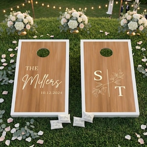 K&ouml;nnte beinhalten: Zwei Cornhole-Spielbretter aus Holz mit wei&szlig;en R&auml;ndern, personalisiert f&uuml;r eine Hochzeit. Auf einem Brett steht "The Millers 10.12.2024", auf dem anderen die Initialen "S T" mit floralen Akzenten. Kleine Beanbags sind sichtbar.