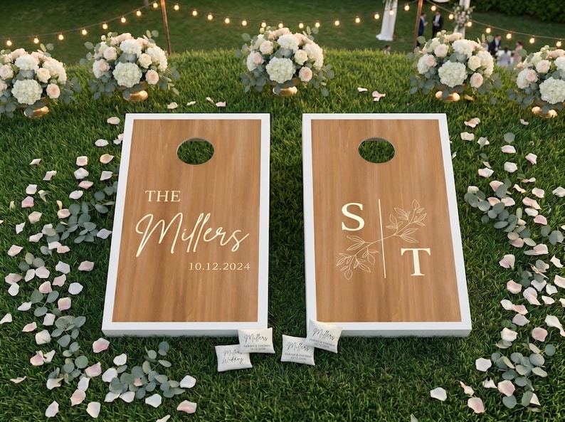 K&ouml;nnte beinhalten: Zwei Cornhole-Spielbretter aus Holz mit wei&szlig;en R&auml;ndern, personalisiert f&uuml;r eine Hochzeit. Auf einem Brett steht "The Millers 10.12.2024", auf dem anderen die Initialen "S T" mit floralen Akzenten. Kleine Beanbags sind sichtbar.