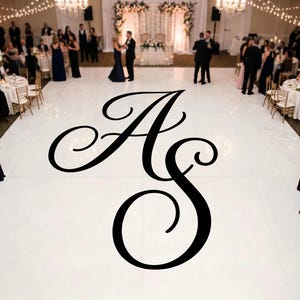 Könnte beinhalten: Ein großes, schwarzes Monogramm in Schreibschrift mit den Buchstaben AS, zentriert auf einer glänzenden weißen Tanzfläche. Die Tanzfläche ist von runden Tischen mit weißen Tischdecken, goldenen Stühlen und Gästen in Abendgarderobe umgeben.