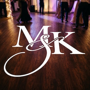 May include: A white monogram projection featuring the letters M and K with an ampersand symbol, displayed on a dark brown wooden dance floor at a wedding reception with blurred guests dancing in the background under warm ambient lighting.