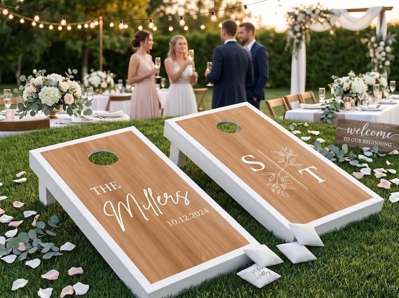 K&ouml;nnte beinhalten: Zwei individuelle Cornhole-Boards mit wei&szlig;en Rahmen und Holzoberfl&auml;chen. Ein Board zeigt "THE Millers" und das Datum "10.12.2024". Das andere Board zeigt die Initialen "S T" mit floralen Akzenten. Kleine wei&szlig;e Beanbags liegen daneben.