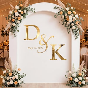 Puede incluir: Un telón de fondo de boda blanco con las iniciales doradas "D & K" y la fecha "17 de mayo de 2027". El telón de fondo está adornado con arreglos florales de rosas blancas y melocotón, vegetación y hierba de la pampa. La escena se desarrolla en un lugar decorado.