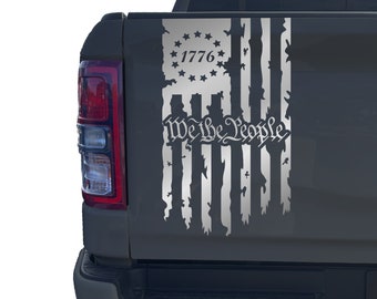 May include: A gray truck tailgate with a distressed American flag decal. The flag has the words "We the People" and "1776" printed on it.