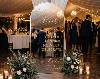 Könnte beinhalten: Ein bogenförmiger Standspiegel mit goldenem Rahmen und goldener Aufschrift: Hello Friends, YOU LOOK STUNNING. NOW LET'S CELEBRATE. xo. Emily & Jack. Der Spiegel reflektiert eine Hochzeitsfeier mit Gästen und ist von Blumenarrangements umgeben.