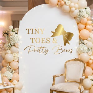 May include: A white arched backdrop featuring the gold text Tiny Toes & Pretty Bows with a gold bow graphic. The arch is decorated with a large balloon garland in shades of cream and peach, accented with white flowers and small fairy lights.