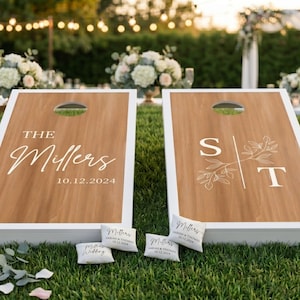 K&ouml;nnte beinhalten: Zwei personalisierte Cornhole-Boards mit wei&szlig;em Rand und Holzmaserung. Ein Board zeigt "The Millers" und das Datum "10.12.2024". Das andere Board hat die Initialen "S" und "T" mit floralen Akzenten. Kleine personalisierte Kissen liegen herum.