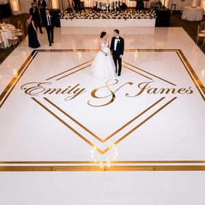Puede incluir: Una gran pista de baile cuadrada de color blanco con un borde geométrico en forma de diamante dorado y los nombres Emily & James impresos en una fuente cursiva dorada en el centro. La pista está en un salón con mesas redondas y flores.
