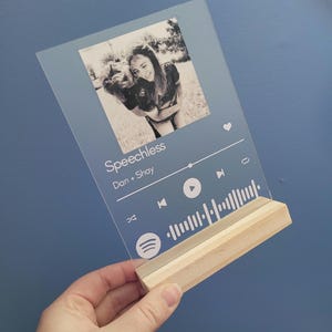 Puede incluir: Una placa de acrílico transparente con una foto en blanco y negro de dos personas, un icono de reproductor de música y el texto "Speechless" y "Dan + Shay". La placa tiene un soporte de madera.