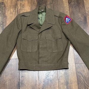 May include: Olive green military jacket with a notched collar, two chest pockets, and a patch on the upper left sleeve. The jacket has long sleeves and a button closure. The jacket is made of a durable fabric.