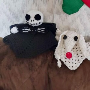 May include: Crocheted Jack Skellington and Zero dolls from The Nightmare Before Christmas. Jack is in a black outfit with a white head and Zero is white with a red nose. The dolls are handmade and sit on a brown surface.