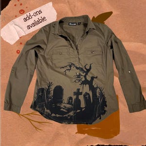 Peut inclure: Chemise boutonnée vert olive avec un motif de cimetière noir. La chemise à manches longues a un col, deux poches de poitrine et des manches retroussables. Le texte "add-ons available" est sur une étiquette blanche.